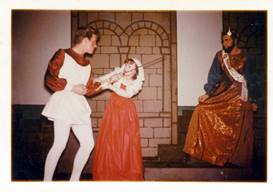 A person in a crown holding a sword to a person in a red dress

Description automatically generated